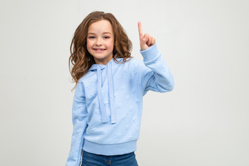 cute teenage girl in blue hoodie shows one finger on a light gray background