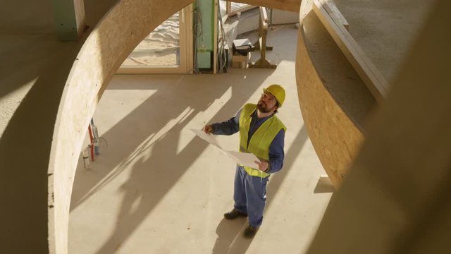 SLOW MOTION: Static engineer looks at his floor plans and observes the prefabricated house under construction. Young Caucasian architect looks at construction plans in a cross-laminated timber house.
