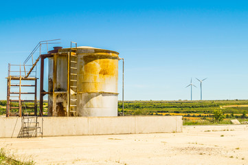 Old oil storage tank with wind turbines in the background on sun