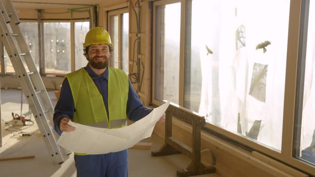 SLOW MOTION, CLOSE UP, PORTRAIT, LENS FLARE: Construction site manager smiles and looks into camera while holding his floor plans. Architect is happy with how the prefabricated house in being built.