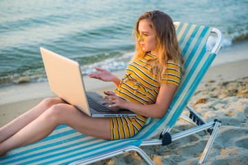 Fit young woman sitting on deck chair at sea view beach using laptop. Female freelance programmer in chaise-long lounge working coding surfing on notebook computer, blank screen. Remote work concept.