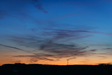 Strommast Freileitung als Silhouette in der Abendämmerung blau zu orange mit Leuchten am Horizont