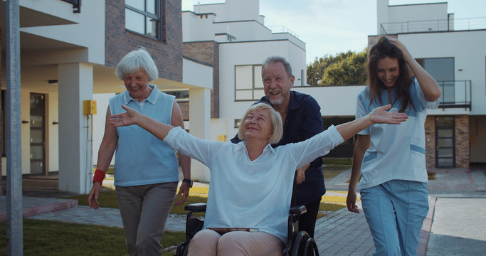 Family of happy disabled woman riding in wheelchair with husband girlfriend and young pretty nurse in the street. Happy moments. Concept of help for elderly.