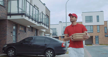 Attractive young delivery man in red uniform with hot pizza boxes walking in the street of modern...