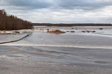 The rocky shore of a large lake in early spring, trees, large stones and melting ice near the shore on a cloudy spring day.