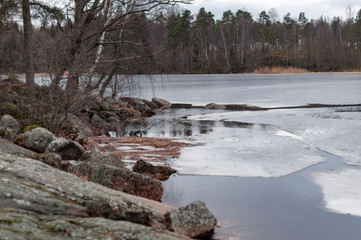The rocky shore of a large lake in early spring, trees, large stones and melting ice near the shore on a cloudy spring day.