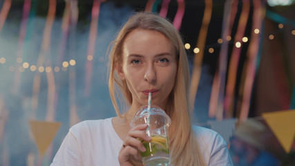 Portrait of beautiful happy blonde girl partying with friends at night. Smiling nice mood young woman drinking cocktail chilling at summer party.