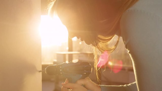 SLOW MOTION, CLOSE UP, LENS FLARE, DOF: Female contractor doing a man's job by using a power drill fasten a wall panel at sunrise. Beautiful Caucasian woman screws small bolts into a gypsum wall board