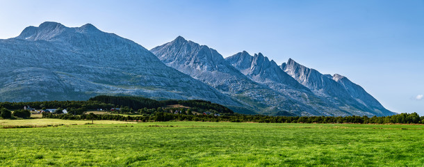 Panoramic view at five of Seven Sisters Mountain range on Sandnessjoen, ideally green grass, small Norwegian village and mountains beside the field. Shinny sunny day, blue skies, Norway, copy space - obrazy, fototapety, plakaty