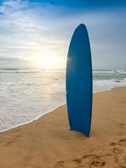 Beautfiul photo of blue surboard standing on the sandy ocean beach at sunset