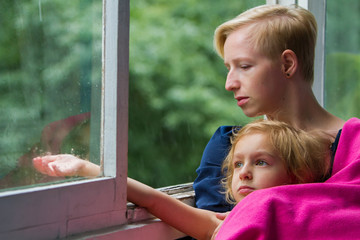 Mother and daughter are sitting by the open window during rain, mother is holding her daughter in a blanket on herself, daughter is sticking her hand out of the window and catching raindrops.