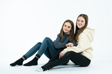 Two cute brunette twins in casual hoodies sitting on the ground and looking at the camera isolated over white background