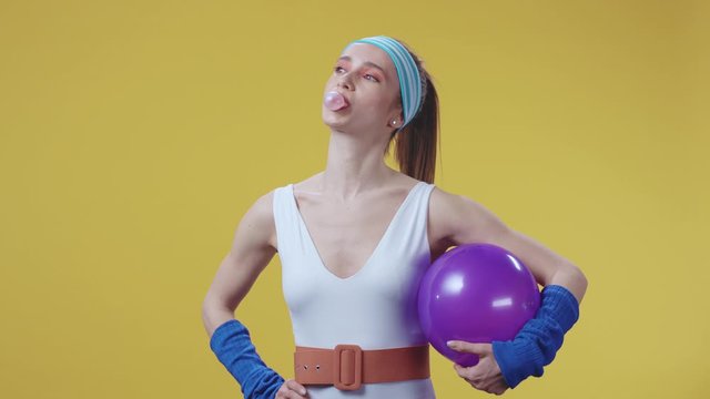 Confident cute retro fitness woman holds a sport gym ball and chewing bubble gum laughing smiling at camera on yellow background.