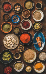 Flat-lay of various spices in bowls and plates over rusty background, top view. Black pepper, allspice, cloves, thyme, cumin, sesame, star anise, cardamom, smoked salt, ginger, cinnamon, rosemary, bay