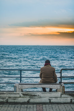 Lonely man sitting alone on the beach at sunset looking away, a ship is pulling away from the horizon