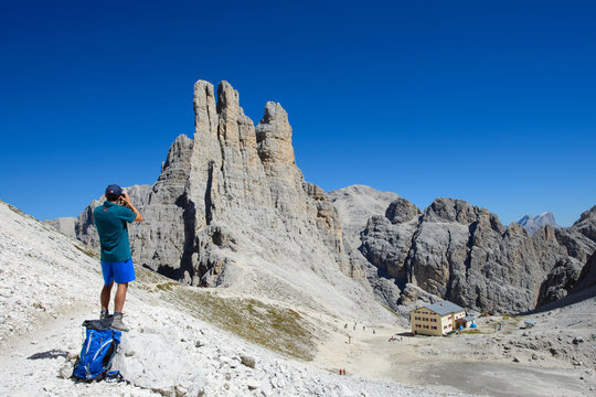 Paesaggio di alta montagna con rifugio  e vette sullo sfondo e persona in primo piano che fotografa le torri del Vaiolet nel parco naturale alpino altezza