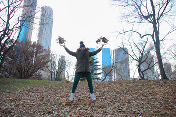 woman in central park jumping, with dry leaves in hands. She is dressed in green pants and jacket. He wears a hat on his head