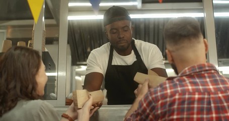 Rear of Caucasian couple standing outdoors at festive food truck cafe window and taking order from African American young vendor. Barman of little street bar giving hot dogs to clients.