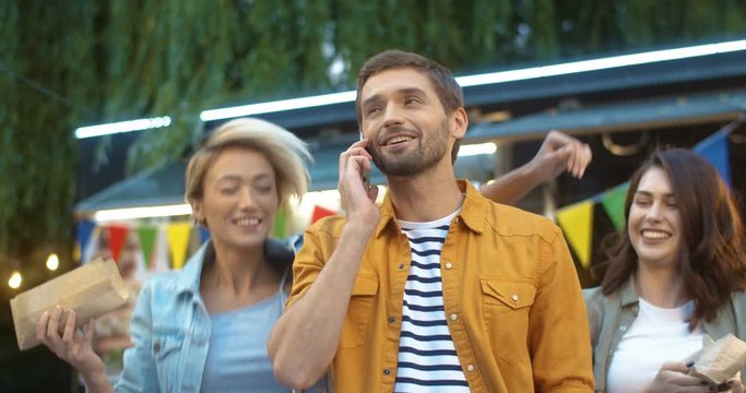 Joyful Caucasian young guy talking on phone with smile outdoor. Two cheerful young women with hot dogs coming to man, hugging and laughing at festive food court in park. Man speaking on cellphone.