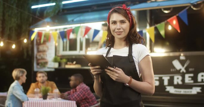 Young pretty Caucasian woman waiter in apron using tablet device in park at food track outdoor. Portrait of beautiful female vendor smiling to camera with truck bar. Girl bartender with computer.