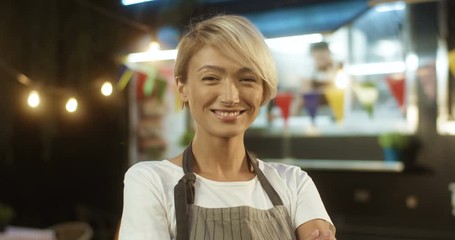 Close up of beautiful joyful young woman in apron with fair short hair smiling at festive food track in evening. Portrait of female Caucasian waiter or vendor at small fair outdoor bar.