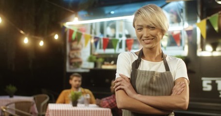 Happy beautiful joyful young woman in apron with blonde short hair smiling at festive food track in evening. Portrait of female Caucasian waiter or vendor at small fair outdoor bar.