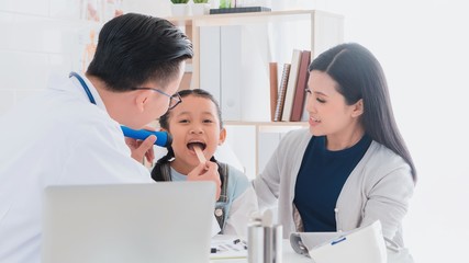 Fototapeta premium Professional doctor wearing white coat using tongue depressor to examine kid patient with her mother in hospital background.Concept of disease treatment and health care in hospitals.