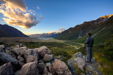 A man wearing a black dress, standing on a rock at the top of the mountain, uses a tripod and a camera to take a panoramic view in the morning. Blue skies and beautiful clouds.