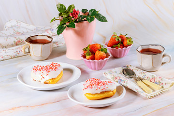 Two sweet and pink heart shaped donut with white glaze and sprinkles on party table with tea, strawberries and rose.