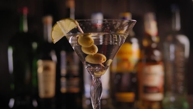 Close-up of a cool cocktail with vermouth vodka or gin with green olives and lime stands on a rotating surface against the background of a bar with bottles. Slow motion.