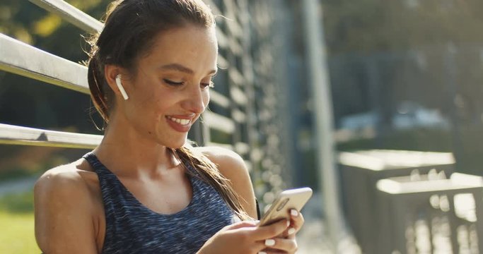 Pretty smiled Caucasian woman in sporty style and headphones scrolling and tapping on smartphone on summer day. Beautiful joyful young girl texting mesage on phone as chatting at sport court.