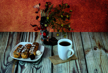 A mug of tea on a napkin, a plate with three eclairs and a glass vase with a herbarium on a wooden...