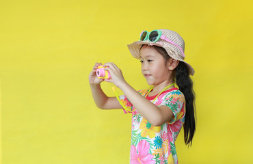 Attractive asian little girl in floral pattern summer dress and hat with sunglasses taking photo by toy camera isolated on yellow background. Fashion of Holiday and summer concept.