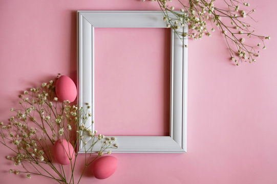 Easter pink eggs and white gypsophila flowers are arranged on either side of a white photo frame on a pink background. Flat lay. The concept of a happy Easter and spring holidays. Copy space.