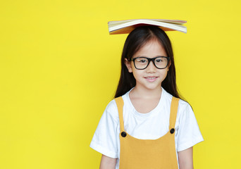Smiling asian little kid girl wearing glasses with book on head and looking camera isolated on...