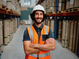 Portrait of cheerful supervisor in a warehouse for delivering and transporting industrial goods wearing white helmet and orange vest uniform standing with folded arms in aisle - obrazy, fototapety, plakaty