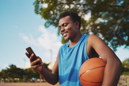 Portrait Of A Smiling Young Male Basketball Player Holding Ball In Hand Texting Messages On His Smart Phone At Park