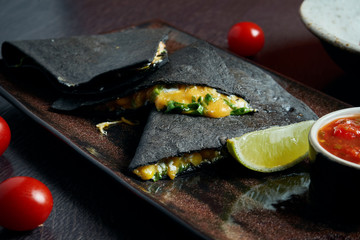 Appetizing baked quesadilla with 4 types of cheese in black tortilla on a ceramic plate on a black background. Modern mexican cuisine.