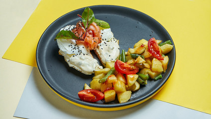 Classic continental breakfast - toasts with cream cheese, salmon and poached egg and a side dish of baked potatoes with tomatoes on a black plate on colored backgrounds
