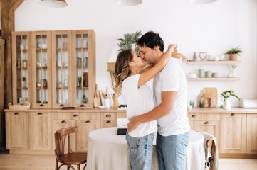 The hugging attractive happy couple in white t-shirts kissing at home in the kitchen