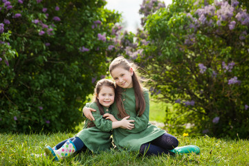 Fototapeta premium Two sisters in green linen dress have fun in the park with blooming lilacs, enjoy spring and warmth
