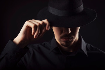 Male with goatee wearing black shirt touching his fedora hat tipped over eyes, dark background