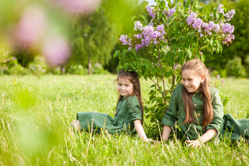 Two sisters in green linen dress have fun in the park with blooming lilacs, enjoy spring and warmth