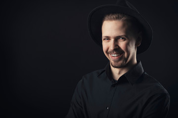 Portrait of smiling guy with chintuft wearing black shirt and fedora hat looking at camera, isolated