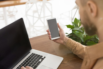 Young man with beard is sitting in cafe, typing on laptop checking his smartphone, multitasking concept, blogging