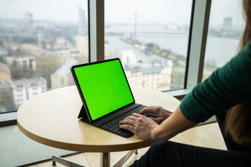 Woman stands by table and watching and typing on professional Digital Tablet Computer with keyboard cover. Background big panoramic windows and city view. Modern interior.