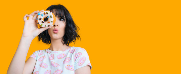 Charming brunette girl looking through a donut is feeling amazed posing on a yellow background with...