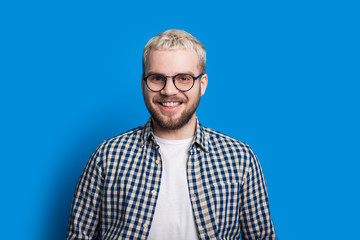 Handsome boy with blonde hair and nice beard is looking through his eyeglasses while posing confidently on blue background