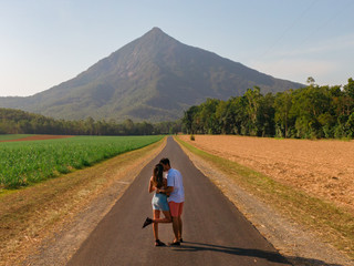 Fototapeta premium Couple with mountain landscape & fields. Sugar cane field. Dramatic DRONE aerial view of fields, trees, green forest, farm, mountains & road. Romantic shot in Walsh's Pyramid, Cairns, Australia.