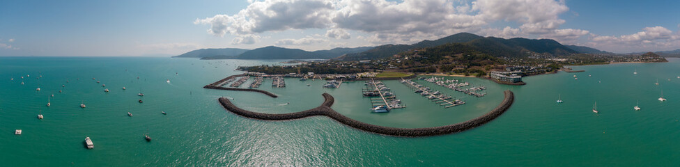Fototapeta na wymiar Panoramic marina town aerial. Airlie beach waterfront aerial view. Dramatic DRONE view from above. Marina town with yachts and boats in sea water. Mountain landscape background. Whitsundays Islands